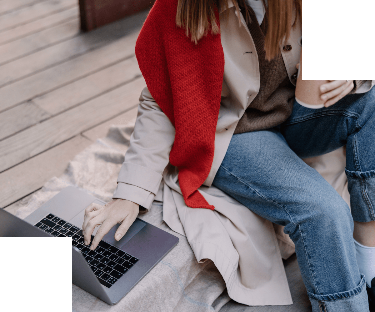 Person Wearing A Beige Trench Coat, Blue Jeans, And A Red Scarf Is Sitting On A Blanket Outdoors, Using A Laptop And Holding A White Cup.